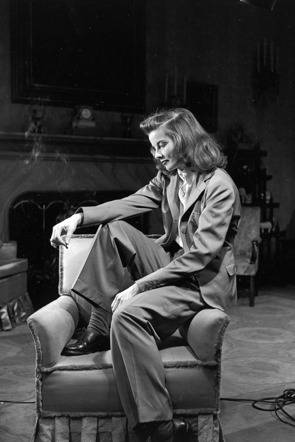 Portrait of actress Katharine Hepburn sitting on arm of chair smoking.