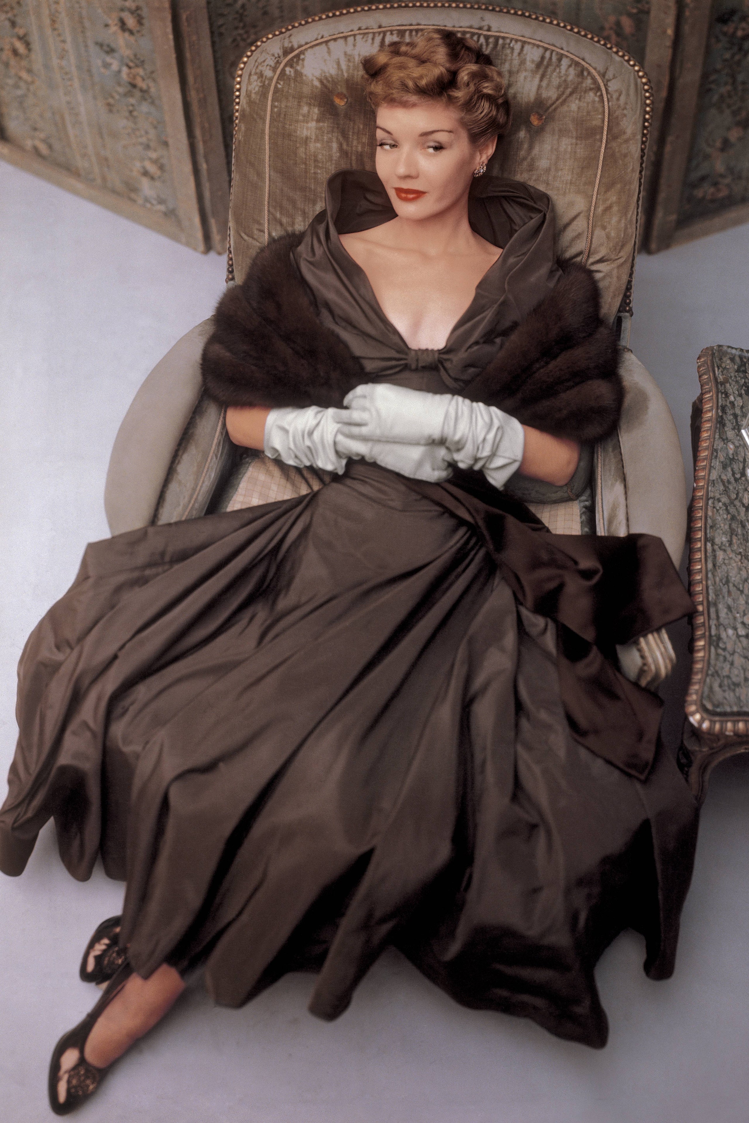 Model seated in a taupe velvet chair wearing a madetoorder taupe taffeta faille evening dress with a stand away neckline...