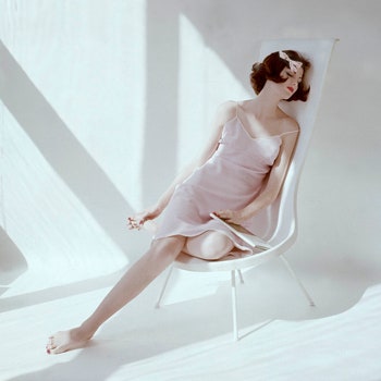 Model sitting in white chair wearing pink and white gingham slip and bow in her hair.