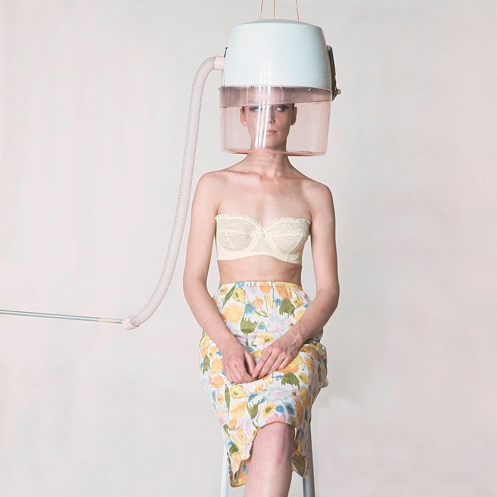 Model with big hair dryer on head wearing lingerie including yellow strapless lace bra and flower print slip .