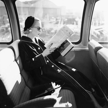 Mrs. William McManus Vogue fashion editor seated in train reading wearing coat sunglasses and hat.