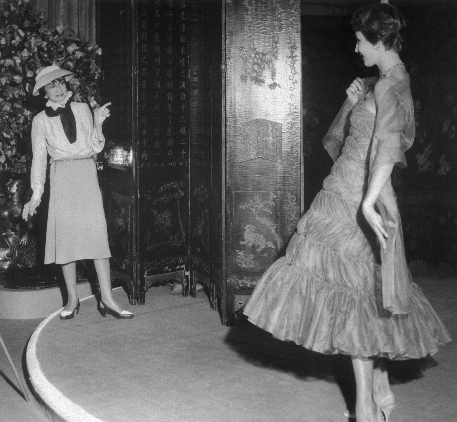 Коко Шанель любуется моделью примеряющей вечернее платье из коричневого шифона с оборками 1957