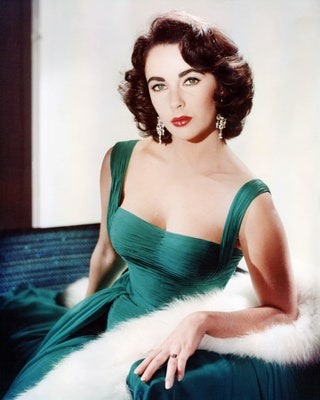 Британская актриса в зеленом платье без рукавов с глубоким вырезом и белой меховой накидкой на подлокотнике кресла около...