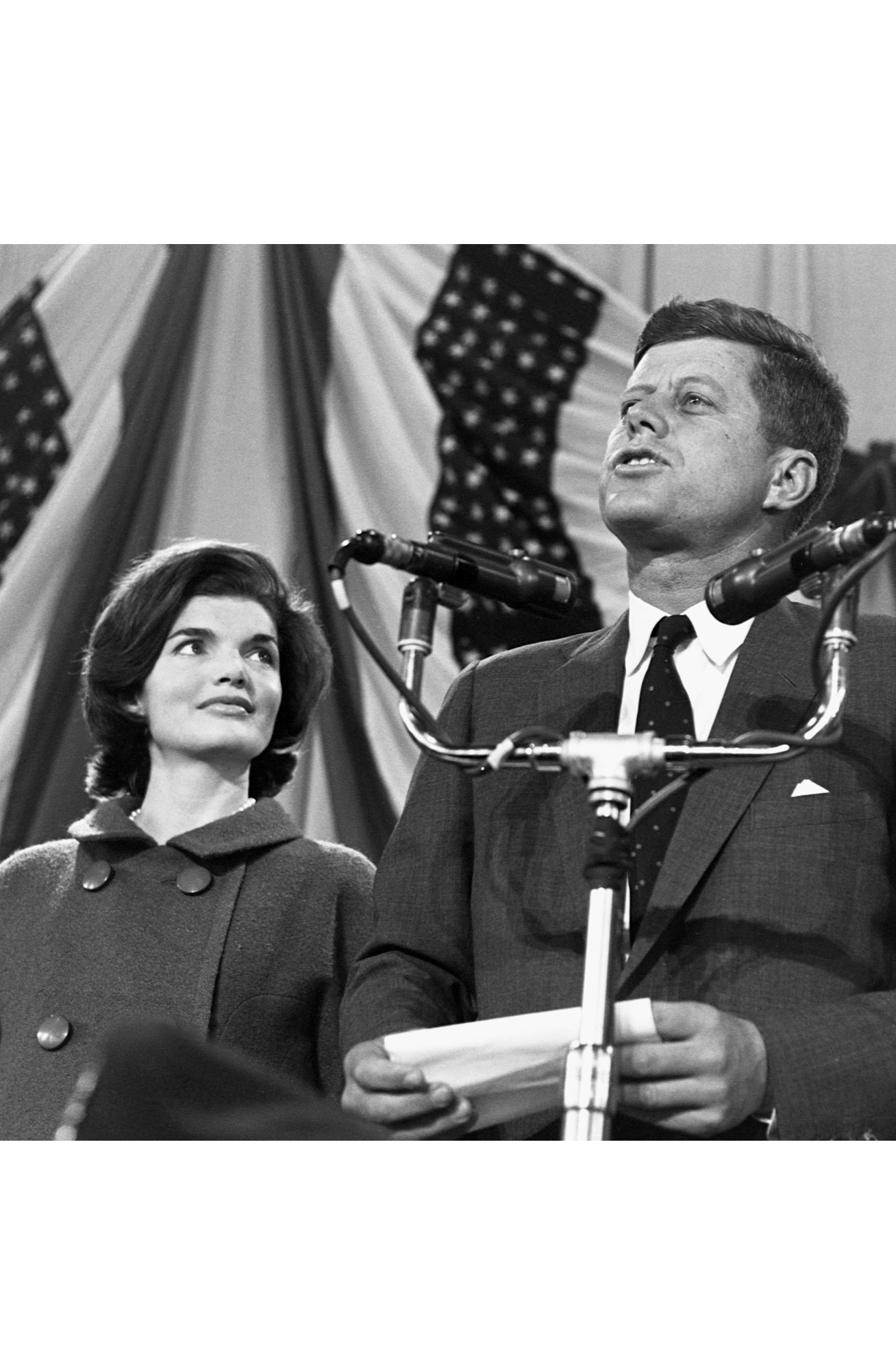 Jacqueline Kennedy watches as husband John F. Kennedy speaks into several microphones in front of a back drop of flags....