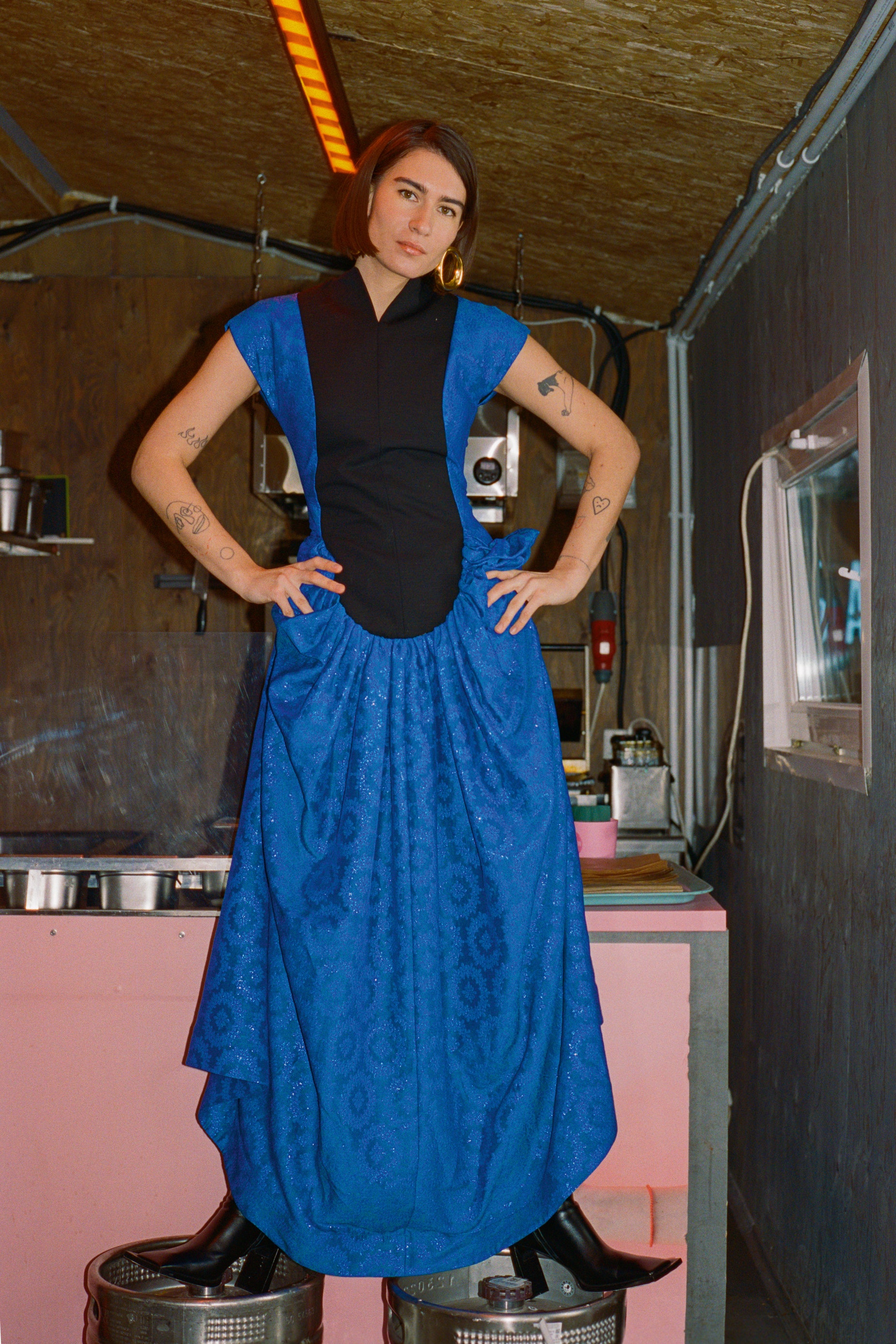 Nick with hand on hip wearing blue coloured silk dress with lurex earrings and black leather boots