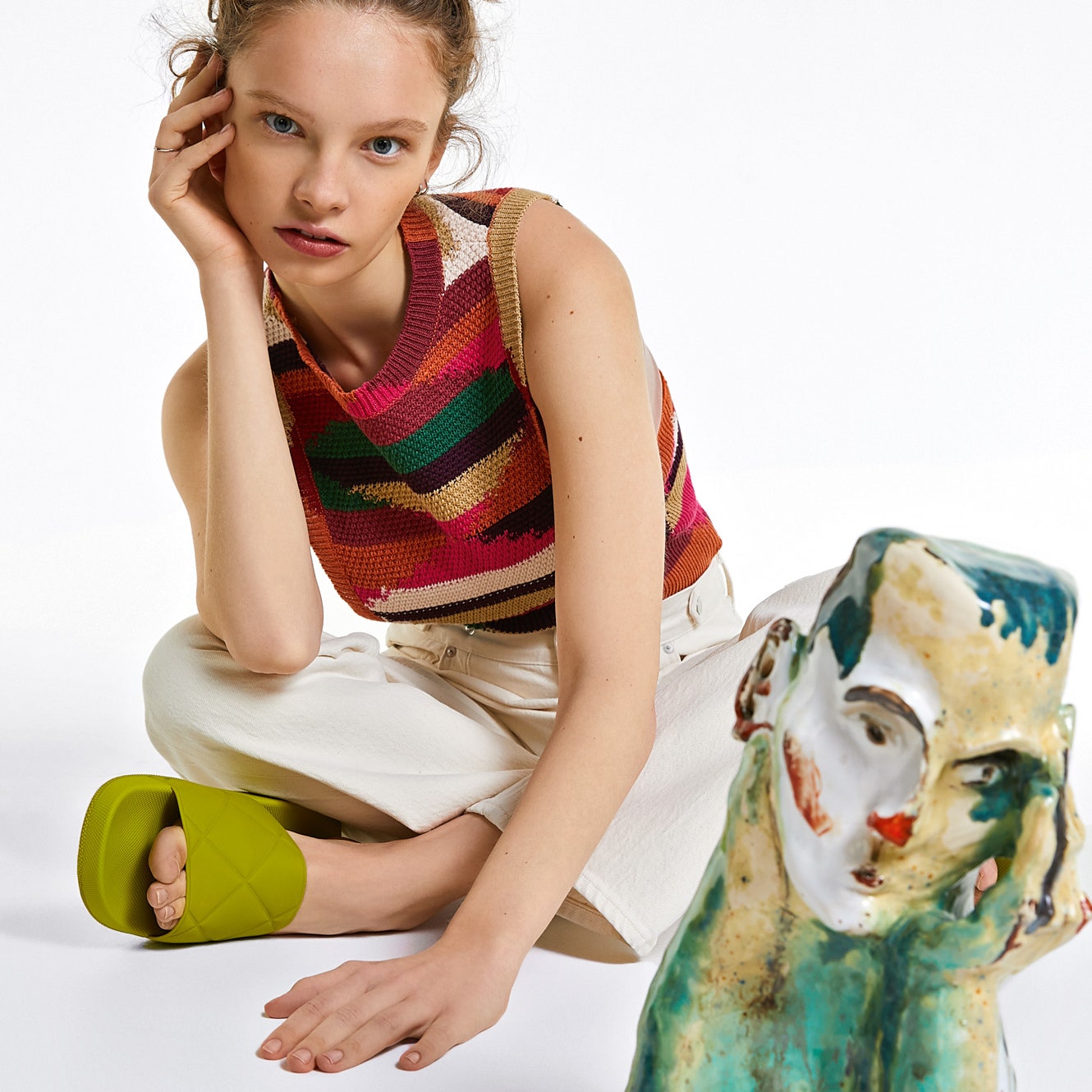 Model wearing striped top white jeans and green sliders sitting near sculpture of woman